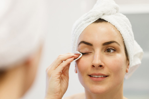 Eine junge Frau mit Handtuchturban auf dem Kopf entfernt mit einem Wattepad und Jojobaöl Make-up von ihrem Auge